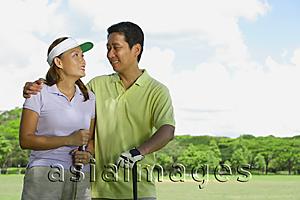 Asia Images Group - Couple standing side by side, holding golf clubs, looking at each other