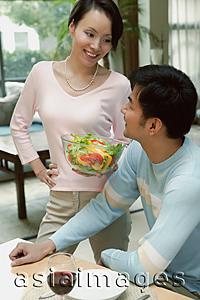 Asia Images Group - Man sitting at dining table looking at woman standing next to him holding bowl of salad