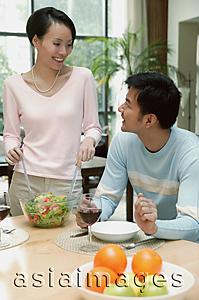 Asia Images Group - Man sitting at dining table, woman standing next to him tossing a salad