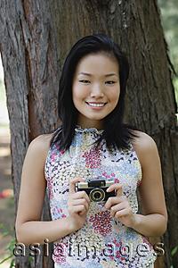 Asia Images Group - Young woman standing next to tree trunk, holding a camera, smiling