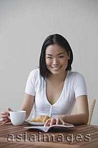 Asia Images Group - Young woman having breakfast and reading a book, smiling at camera