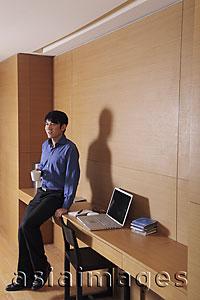 Asia Images Group - Young man sitting on the side of his desk drinking coffee