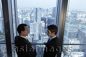 Asia Images Group - Two men talking in front of window with a view of the city of Beijing