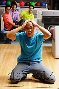 AsiaPix - Man kneeling in bowling alley, hands on head, frowning, people in the background watching