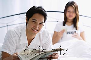 AsiaPix - Couple on bed, man with newspaper, smiling, woman behind him reading book