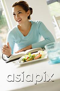 AsiaPix - Young woman sitting at table, salad on table, writing in notebook, looking away