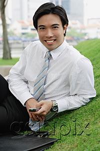 AsiaPix - Businessman sitting on grass in park, holding mobile phone, smiling at camera
