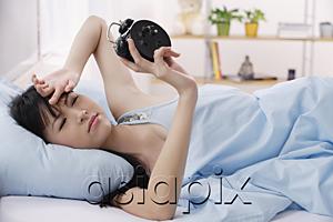 AsiaPix - Young woman lying on bed, frowning at alarm clock