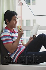AsiaPix - Man sitting on bay window, reading a book, eating a sandwich