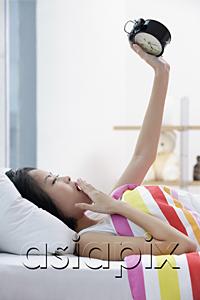 AsiaPix - Young woman lying on bed, looking at alarm clock, hand on mouth