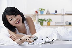 AsiaPix - Young woman lying on bed, holding TV remote control