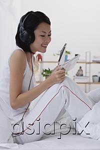 AsiaPix - Young woman sitting on bed, listening to music, smiling
