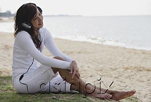 AsiaPix - Young woman sitting on beach, listening to music through headphones