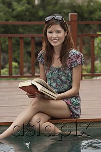 AsiaPix - Woman sitting at the edge of swimming pool with a book, smiling