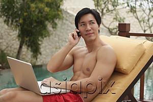 AsiaPix - Man sitting by swimming pool, using laptop and mobile phone