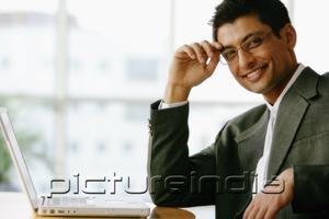 PictureIndia - Man with hand on head, looking at camera, laptop on table