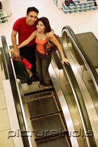 PictureIndia - Young man and woman standing on escalator, looking up at camera