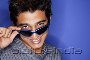 PictureIndia - Man against blue background, adjusting sunglasses, smiling at camera