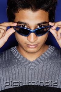 PictureIndia - Man against blue background, adjusting sunglasses, looking at camera