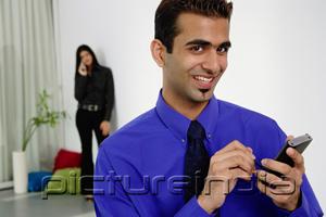 PictureIndia - Businessman using PDA, smiling at camera, woman in the background