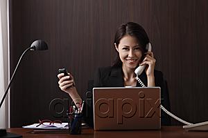 AsiaPix - Woman sitting at her desk smiling and holding her phones