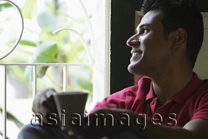 Asia Images Group - Indian man looking out window smiling holding coffee cup