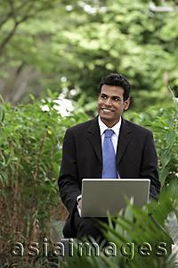 Asia Images Group - Indian man smiling while working on laptop with green plants in foreground and background