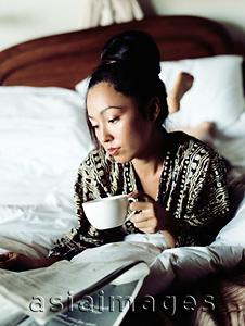 Asia Images Group - Woman in bed reading newspaper, holding cup.