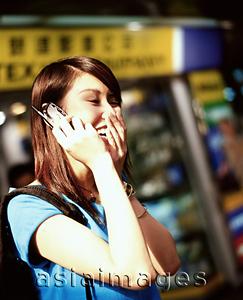 Asia Images Group - Young woman using cellular phone, laughing, shops in background.