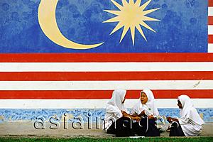 Asia Images Group - Malaysia, Kukup, three Muslim schoolgirls chatting in front of a large Malaysian flag painted on the wall behind them.
