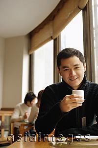 Asia Images Group - Young man sitting at table holding teacup, looking at camera