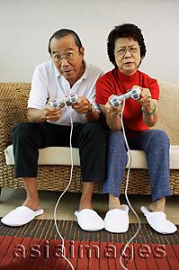 Asia Images Group - Mature couple sitting on sofa playing video games, frowning