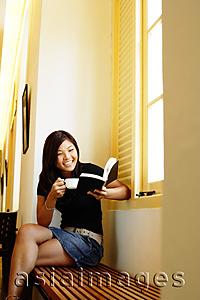 Asia Images Group - Young woman sitting, reading a book and holding cup of coffee