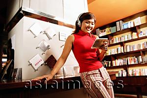 Asia Images Group - Young woman listening to music, smiling, leaning against counter