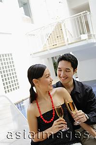 Asia Images Group - Couple sitting by poolside, looking at each other, holding champagne glasses