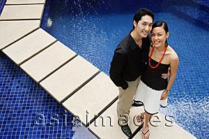 Asia Images Group - Couple standing by swimming pool, looking up at camera
