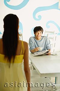 Asia Images Group - Man sitting at table with newspaper and cup, smiling at woman standing in front of him