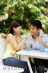 Asia Images Group - Couple sitting side by side, eating, woman feeding man