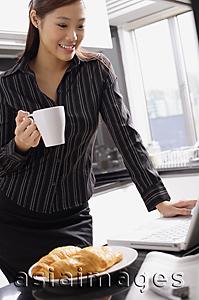 Asia Images Group - Female executive standing in kitchen, holding mug, looking at laptop