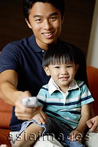 Asia Images Group - Father with young son on lap, holding TV remote control towards camera