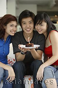 Asia Images Group - Teenagers sitting side by side, smiling at camera, boy in the middle holding plate with a cake and candle on it