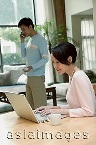 Asia Images Group - Couple at home, woman using laptop, man in the background on the phone