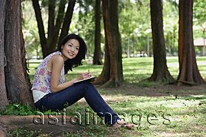 Asia Images Group - Young woman sitting under a tree, smiling, looking away