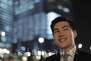 Asia Images Group - Head shot of young man looking up and smiling, lit buildings in background