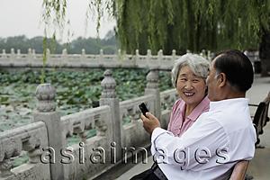 Asia Images Group - Older couple sitting on park bench looking at phone and smiling