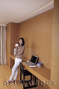 Asia Images Group - Young woman leaning against desk holding coffee