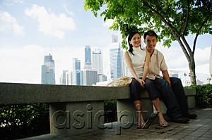 AsiaPix - Young couple sitting on park bench, looking at camera, buildings in the background