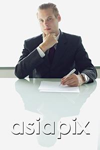 AsiaPix - Businessman sitting at table with pen and paper, looking at camera, hand on chin