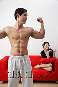 AsiaPix - Couple at home, man in foreground showing off muscles, woman in background, sitting on sofa