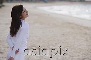 AsiaPix - Young woman standing on beach, hands behind back, looking away